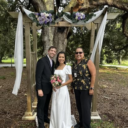 Esther with newlyweds beneath a rustic wooden arbor